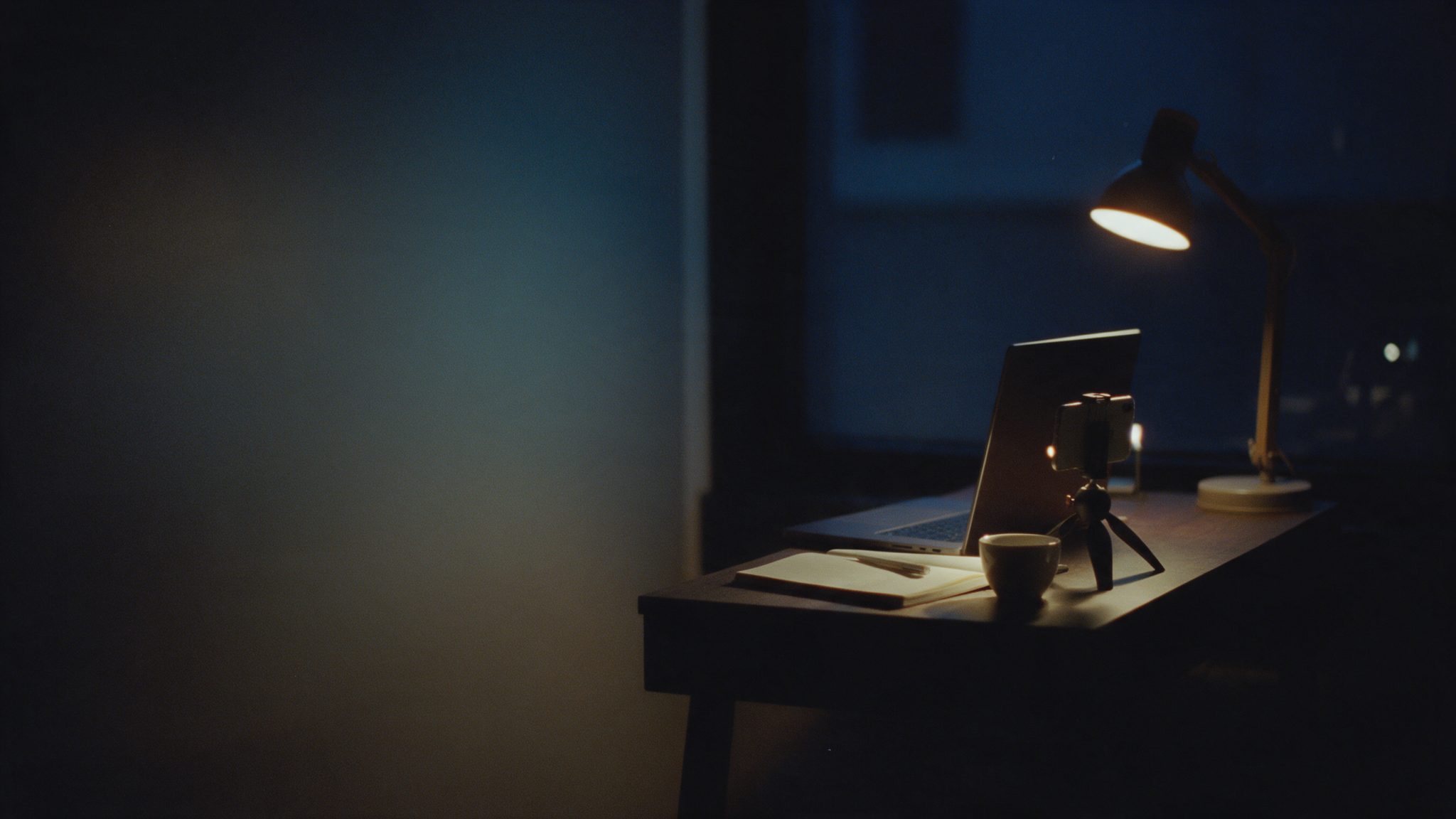 A small worktable at night: a laptop, a phone on a tripod with a small light, a notebook and a lamp.