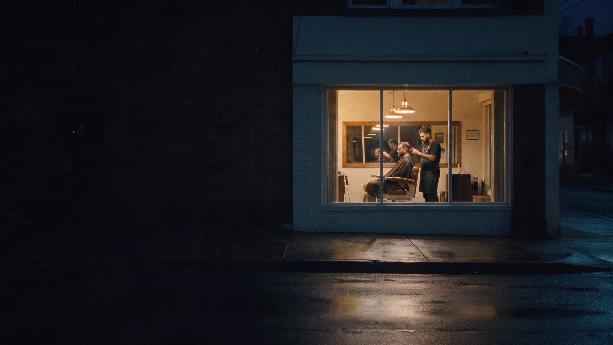 A small shop on a quiet street at night, seen from across the road. Through the lit window, a local business is still at work.