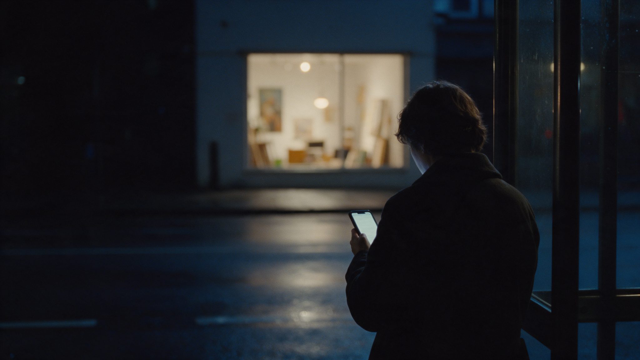 A person at a bus stop at night looks at a phone. Across the street, a small business window is lit.